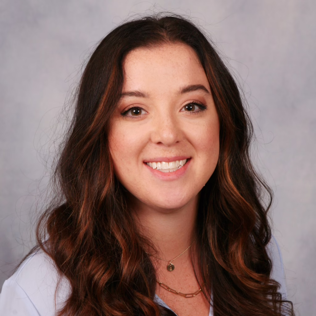 Portrait of a smiling young woman with long wavy brown hair and subtle makeup against a gray studio backdrop, wearing a light blue top and gold necklace(s).
